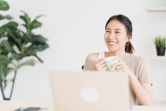 Beautiful young smiling asian woman working on laptop and drinki